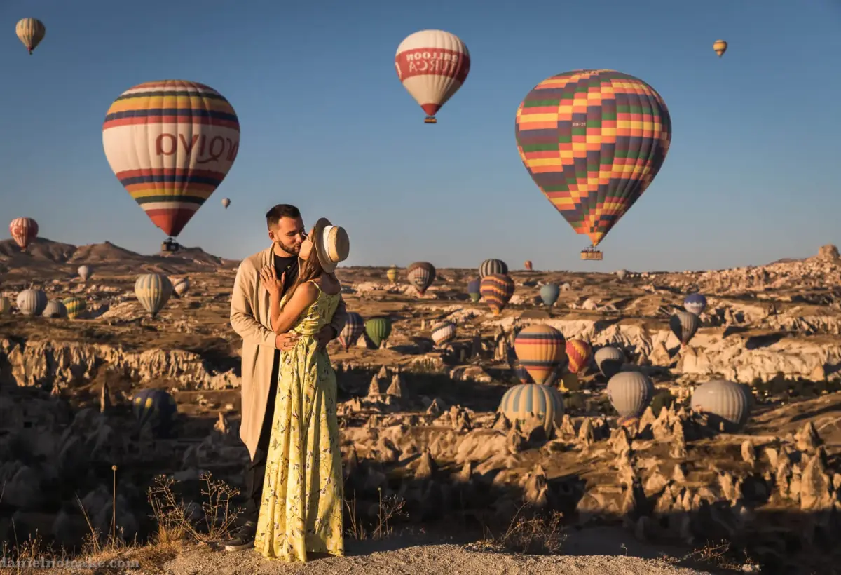 A couple holds each other in a Cappadocia valley with two large striped balloons hovering close behind and dozens more on the horizon.