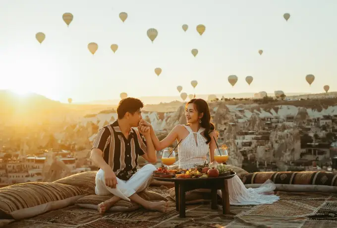 A couple shares breakfast at a low table on a rug spread over the rock in Cappadocia, the sun rising through a sky full of balloons.