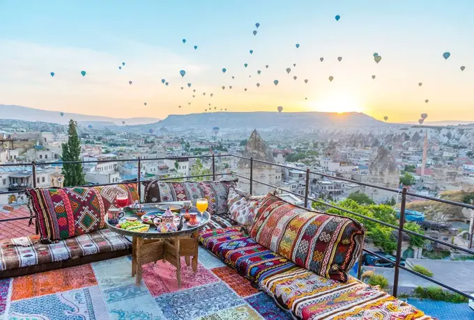 A rooftop terrace in Cappadocia is laid with kilim cushions and a breakfast table as the sun rises over a valley thick with balloons.