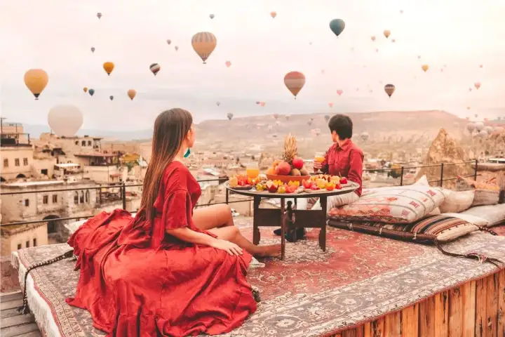 A woman in a flowing red dress sits on a carpeted rooftop in Cappadocia beside a table of fruit, watching balloons rise over the stone town.