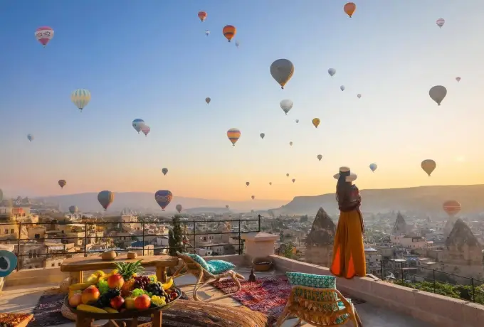 A bowl of fruit sits on a painted terrace table in Cappadocia while a woman at the parapet watches balloons drift across the dawn sky.