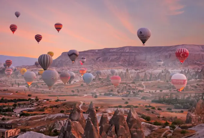 Balloons in every colour crowd the air just above a Cappadocia valley at dawn, pink cloud streaking the sky over the flat-topped ridge behind.