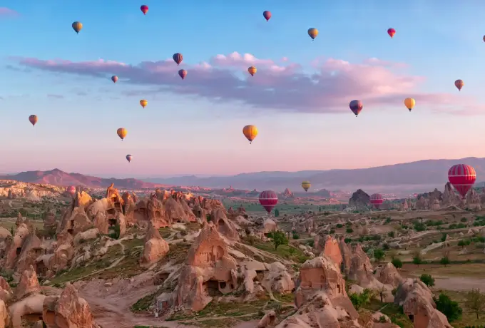 Hot air balloons hang in a pastel pink and blue dawn sky above the eroded rock cones and cultivated valley floor of Cappadocia.