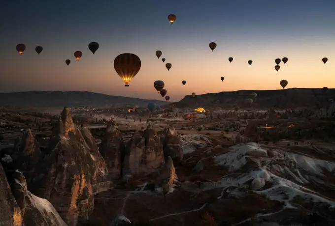 Balloons climb in near darkness before sunrise in Cappadocia, only a thin band of orange light separating them from the black rock valley.