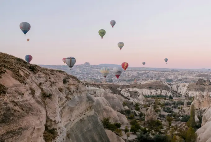 From a high ridge in Cappadocia, hot air balloons float across a deep valley of pale rock and scattered green trees in hazy light.