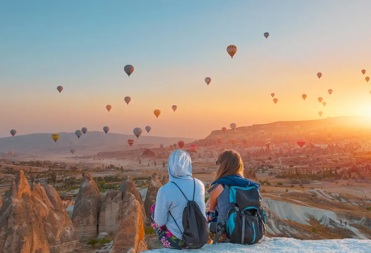 Two travellers with backpacks sit on a bare white rock in Cappadocia, watching hot air balloons climb into the sunrise across the valley.