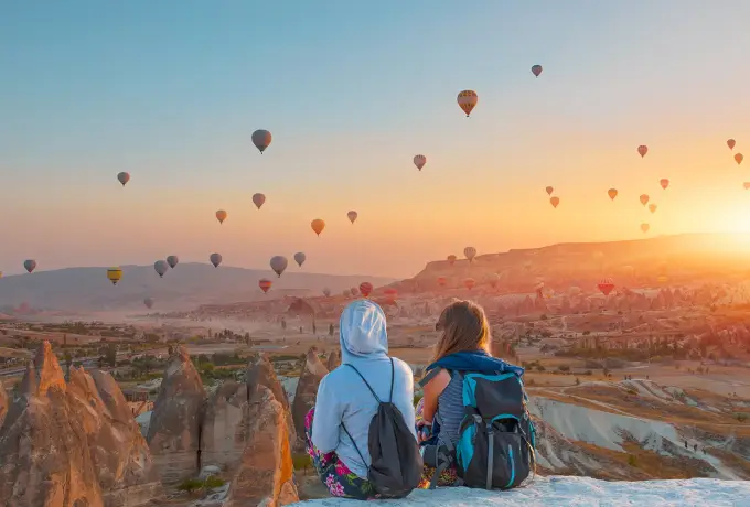 Two travellers with backpacks sit on a bare white rock in Cappadocia, watching hot air balloons climb into the sunrise across the valley.