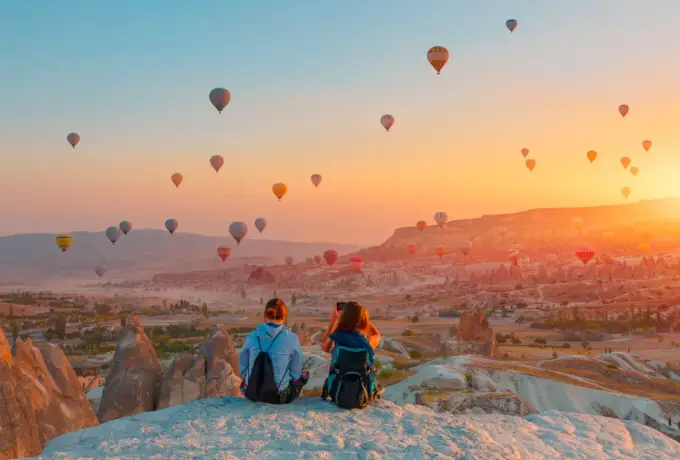 A pair sit close together on a pale rock ridge in Cappadocia as dozens of balloons drift across a blazing sunrise over the valleys.