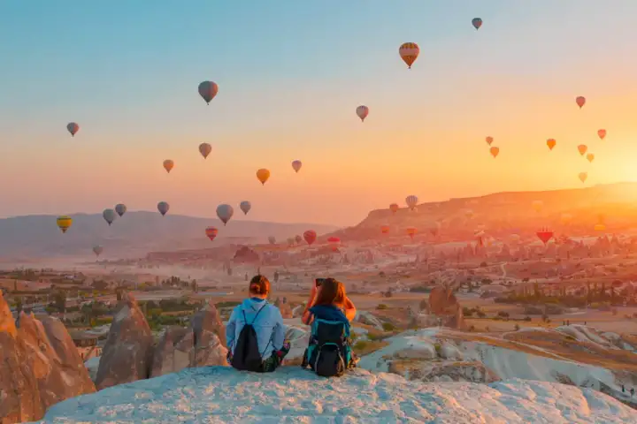A pair sit close together on a pale rock ridge in Cappadocia as dozens of balloons drift across a blazing sunrise over the valleys.