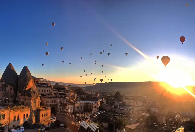 The sun bursts over the ridge behind Goreme in Cappadocia while dozens of balloons climb through the cool blue morning sky.