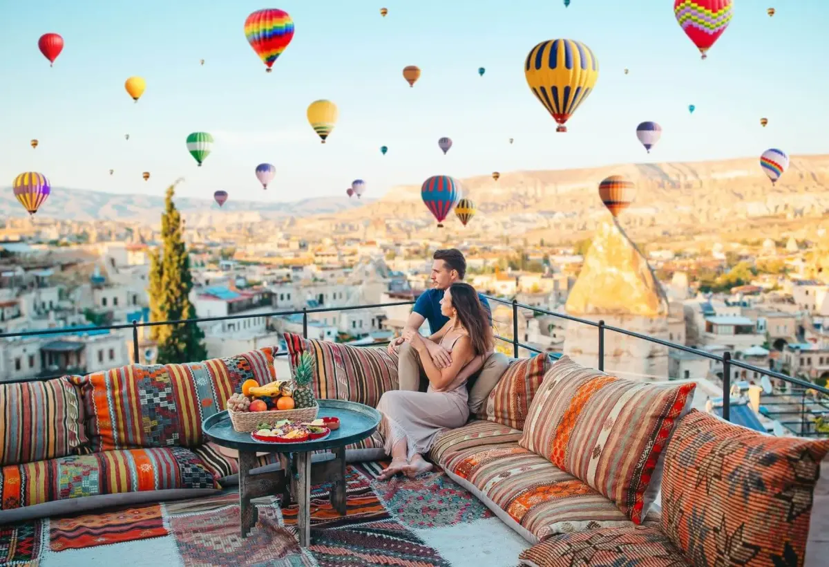 A couple relaxes on a kilim-covered rooftop bench in Cappadocia beneath a sky thick with hot air balloons above the cave town.