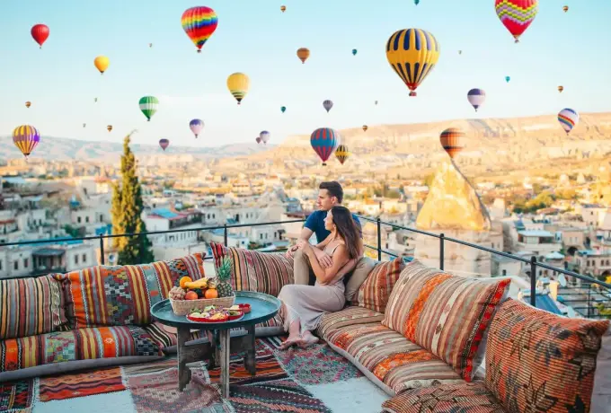 A couple relaxes on a kilim-covered rooftop bench in Cappadocia beneath a sky thick with hot air balloons above the cave town.
