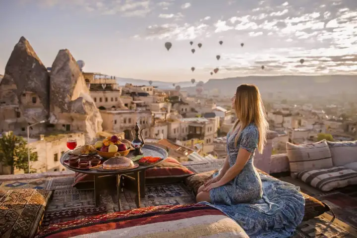 A woman sits on a rug-covered rooftop terrace in Cappadocia beside a laid breakfast table, cave houses and rock cones filling the view behind her.