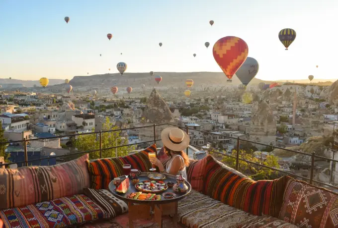 A woman in a wide sun hat sits on a kilim terrace in Cappadocia while a large red and white balloon passes low over the rock town.