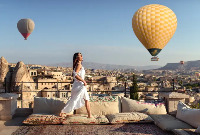 A woman in a flowing white dress stands on a cushioned rooftop in Cappadocia as a large yellow balloon drifts past at close range.