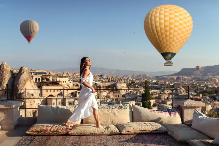 A woman in a flowing white dress stands on a cushioned rooftop in Cappadocia as a large yellow balloon drifts past at close range.