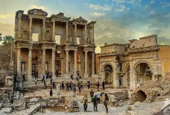 Amber and violet cloud gathers over the Library of Celsus and the arched gate beside it as visitors cross the forecourt at Ephesus.