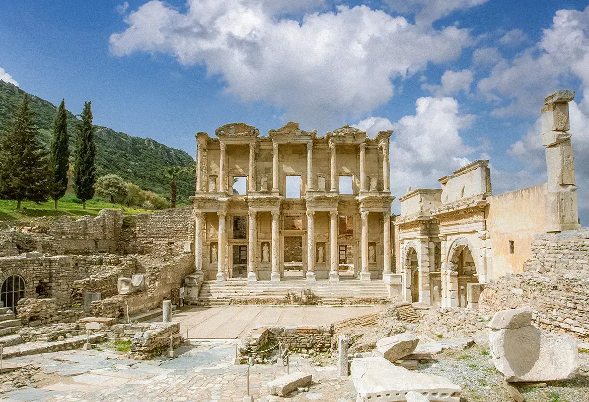 A wide view sets the Library of Celsus at Ephesus between cypress trees and a forested hillside under drifting white cloud.
