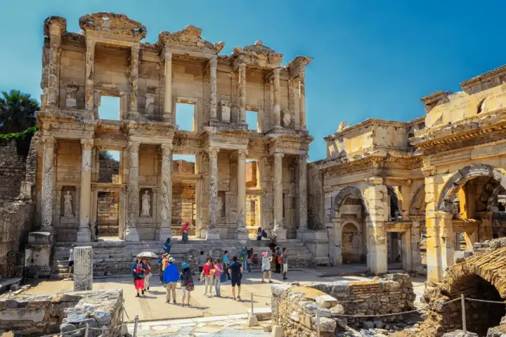 Visitors gather at the base of the Library of Celsus at Ephesus, the arched Roman gate beside it under a clear turquoise sky.