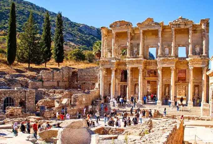 Visitors fill the forecourt of the Library of Celsus at Ephesus on a clear autumn day below a cypress-covered hillside.