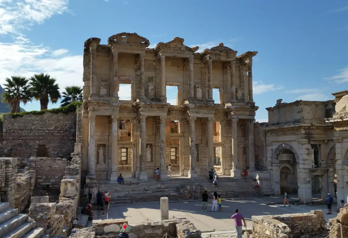 A frontal view of the Library of Celsus at Ephesus under soft midday light, a palm tree rising behind the left of the facade.