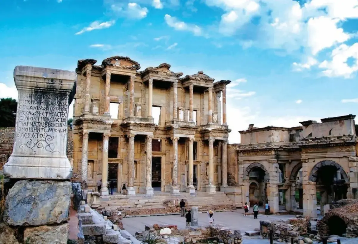 An inscribed marble stele stands in the foreground with the Library of Celsus and the Roman gate rising behind it at Ephesus.