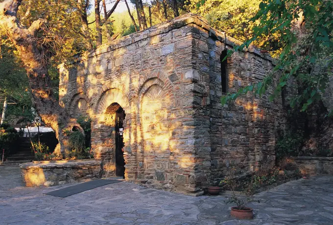 Late afternoon sun lights the stone walls of the House of the Virgin Mary, long tree shadows falling across the paved courtyard.