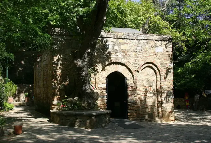 A spreading tree half hides the arched stone doorway of the House of the Virgin Mary in deep summer shade.