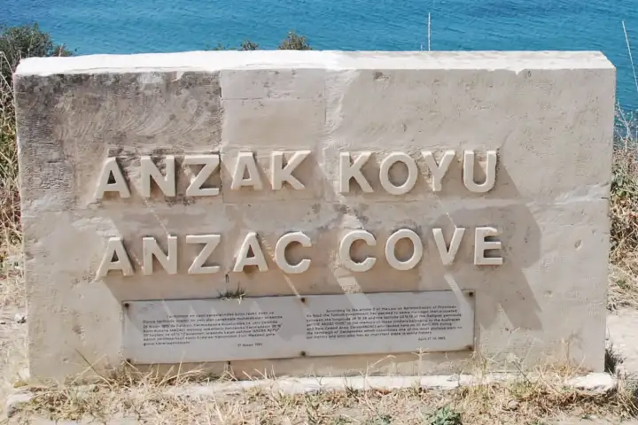 A carved stone marker naming Anzac Cove in Turkish and English stands on the dry grass above the Gallipoli shore.
