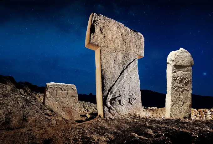 Three T-shaped limestone pillars at Gobeklitepe stand spotlit against a deep blue night sky, animal reliefs visible on their faces.