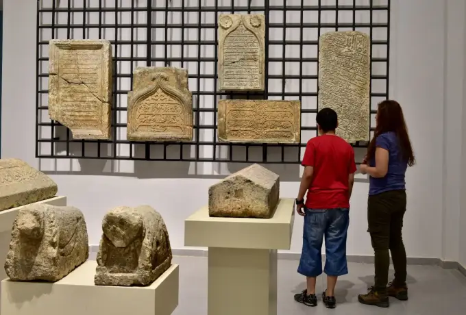 Two visitors study a grid of inscribed stone tablets in the Sanliurfa museum, carved lion tomb markers standing on pedestals nearby.
