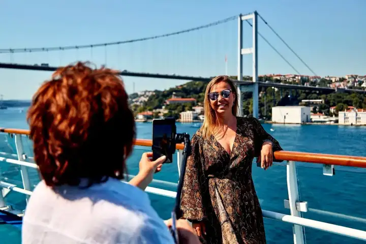 A passenger poses at the rail of a Bosphorus cruise boat in Istanbul while a friend takes the photo, the bridge behind them.