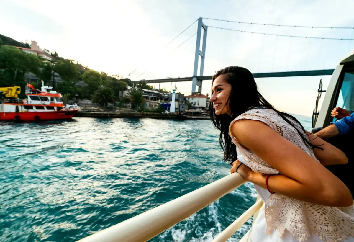 A passenger leans on the rail of a Bosphorus cruise boat in Istanbul, the wake churning turquoise below the bridge.