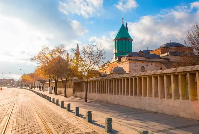 A long colonnaded wall runs beside a wide paved boulevard in Konya with the turquoise dome of the Mevlana Museum above the roofline.
