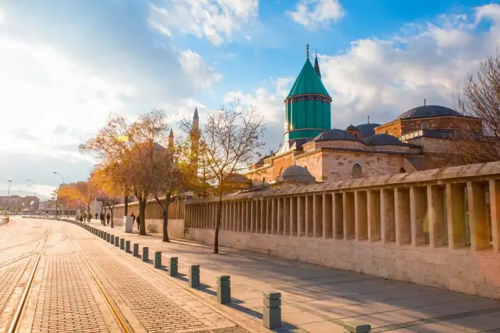 A long colonnaded wall runs beside a wide paved boulevard in Konya with the turquoise dome of the Mevlana Museum above the roofline.