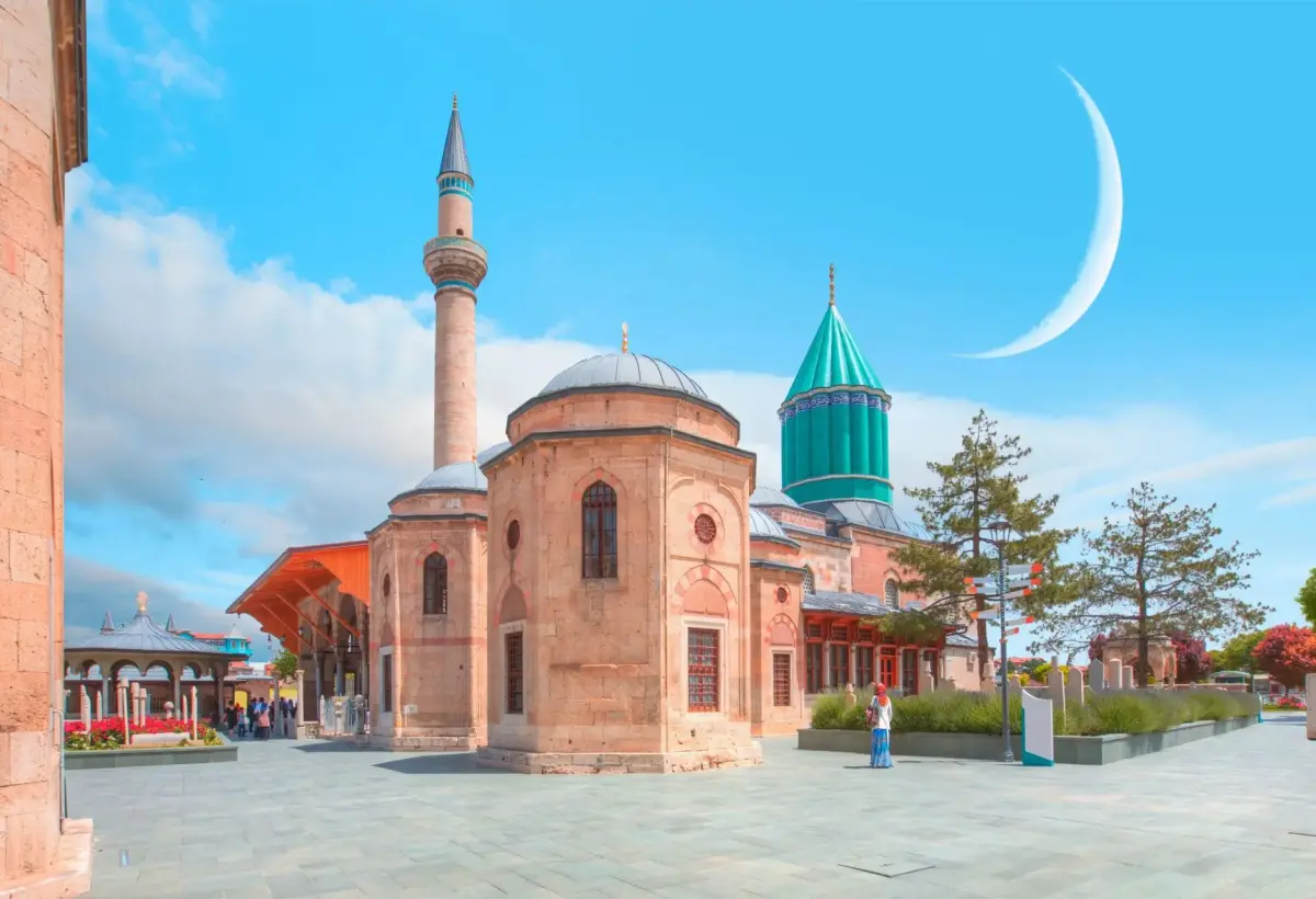 A crescent moon hangs in the bright sky above the octagonal mosque, minaret and turquoise dome of the Mevlana complex in Konya.