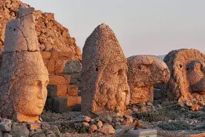 Colossal stone heads and a pointed crown rest on the terrace of Mount Nemrut, lit warm amber by the setting sun.