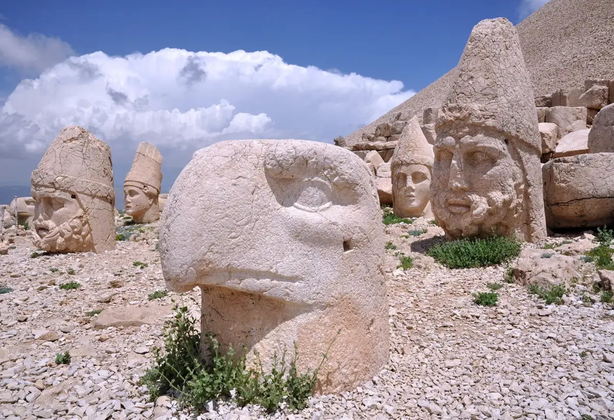 The stone eagle head sits in the foreground of the Nemrut terrace beside a calm-featured deity face under white cloud.