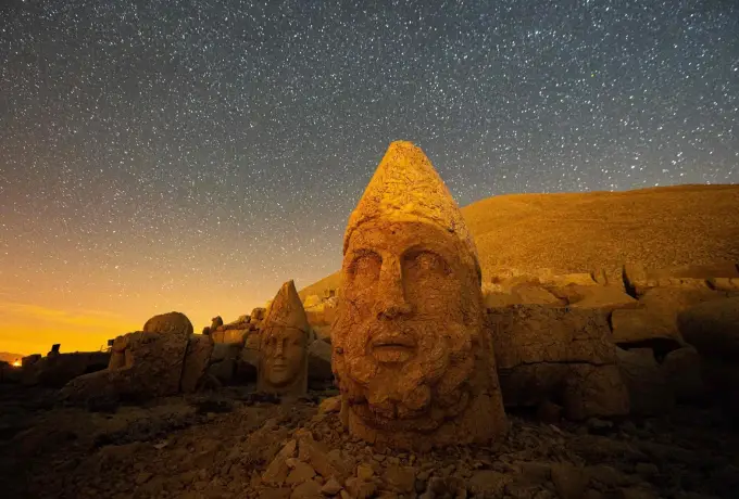 A cone-crowned stone head glows warm gold on the Nemrut terrace beneath a sky thick with stars.