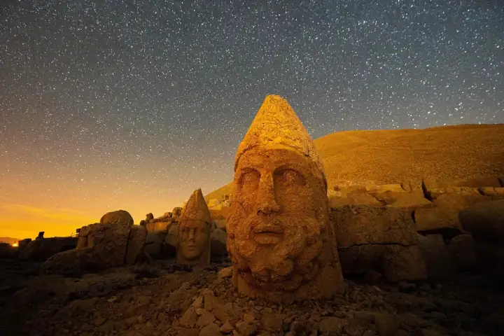 A cone-crowned stone head glows warm gold on the Nemrut terrace beneath a sky thick with stars.