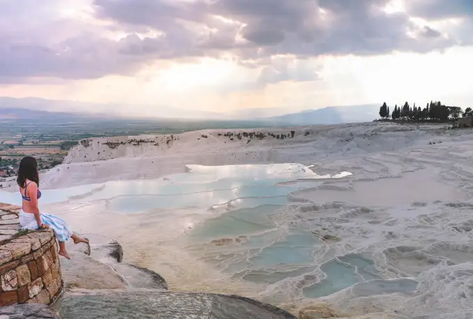 A visitor sits on a ledge above the wide white travertine slope at Pamukkale as evening light spreads across the valley.
