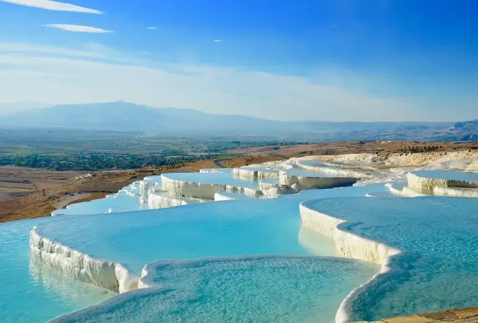 Bright turquoise pools sit in white travertine basins at Pamukkale above green farmland and a distant snow-capped peak.