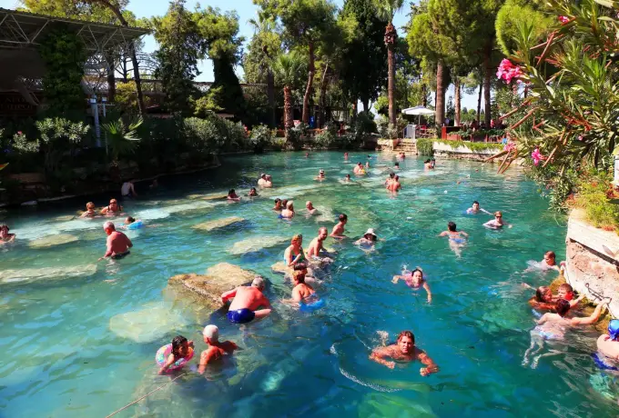 Bathers fill the open-air Cleopatra pool at Pamukkale, surrounded by palms and dense greenery over sunken marble ruins.