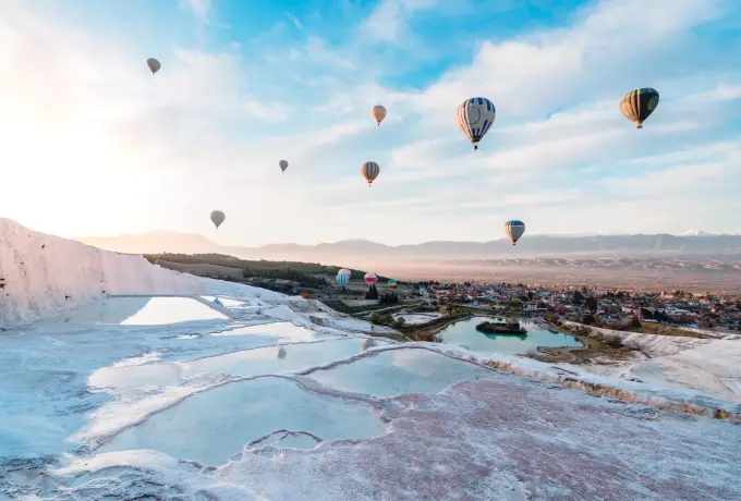 Hot air balloons drift above the white travertine hillside at Pamukkale with the town and valley spread out below.
