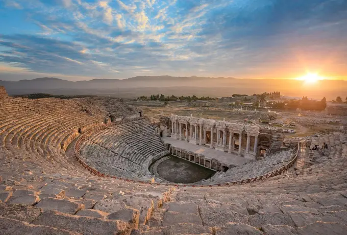 The sun rises behind the Hierapolis theatre at Pamukkale, lighting its seating tiers and the plain beyond.