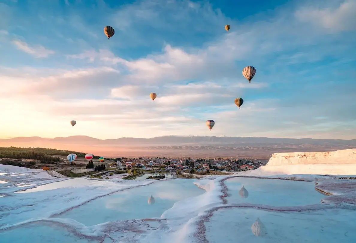 Hot air balloons rise into a pale dawn sky above the white terraces of Pamukkale, the pools mirroring the soft colour.