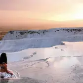 A woman walks alone across the wet white travertine at Pamukkale as the sun sets over the wide plain beyond.