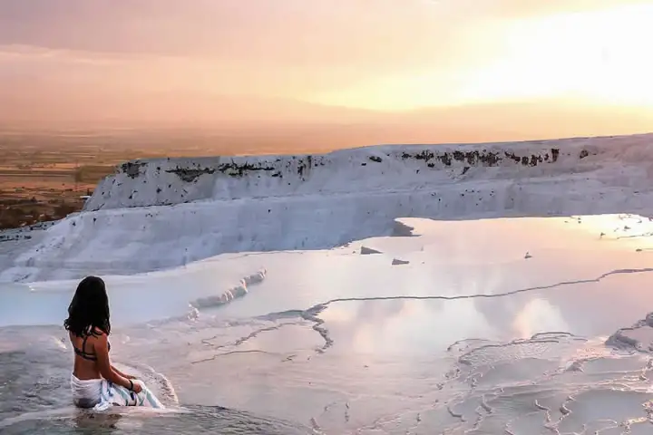 A woman walks alone across the wet white travertine at Pamukkale as the sun sets over the wide plain beyond.