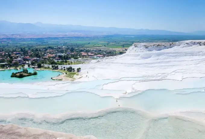 The full white travertine hillside of Pamukkale drops toward the thermal lake and the flat green plain of Denizli.