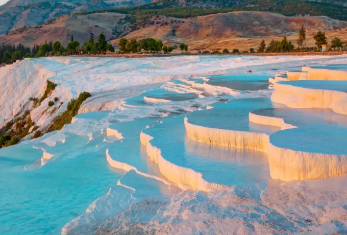 White travertine rims hold turquoise water at Pamukkale below a wooded slope touched with autumn colour.